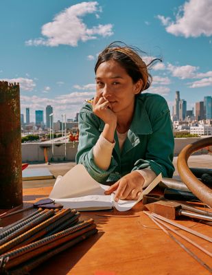 Mujer sentada al aire libre usando un IQOS Iluma i ONE azul con fondo de ciudad.