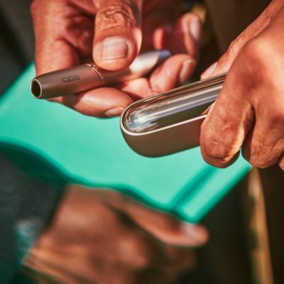 Sunlight shining on a brilliant gold IQOS device someone's right hand, charger in left