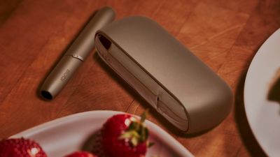 A gold IQOS device and holder next to a bowl of fruit.
