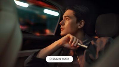 A person is sitting in a car and looking out the window past blurring lights, holding an IQOS ILUMA holder close. At the bottom of the image, there is a white button with the text 'Discover more'.