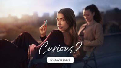 Two people sitting outdoors with a blurred cityscape in the background. One person is holding an IQOS ILUMA holder, and the other person is sitting slightly behind. The text on the image reads: 'Curious?' followed by a button that says, 'Discover more'.