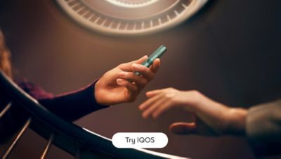 A person is handing a Jade Green-colored IQOS ILUMA device to another person. The background features a circular light fixture, and the text 'Try IQOS' is displayed at the bottom center of the image.