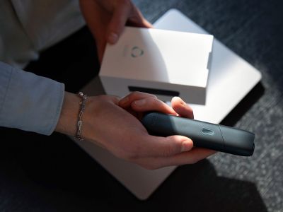 Hand hält IQOS ORIGINALS ONE Slate vor geöffneter Geräte-Verpackung.