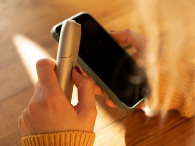 Frau hält IQOS ORIGINALS ONE Silver in linker und Smartphone in rechter Hand.