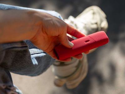 Hand hält IQOS ORIGINALS ONE Scarlet mit roter Schutzhülle vor unscharfem Hintergrund.