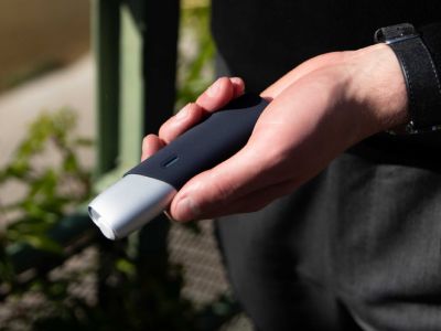 Hand hält IQOS ORIGINALS ONE Silver mit schwarzer Schutzhülle vor unscharfem Hintergrund.