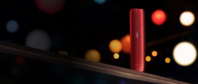 IQOS ORIGINALS ONE Scarlet vor dunklem Hintergrund mit bunten Lichtern.
