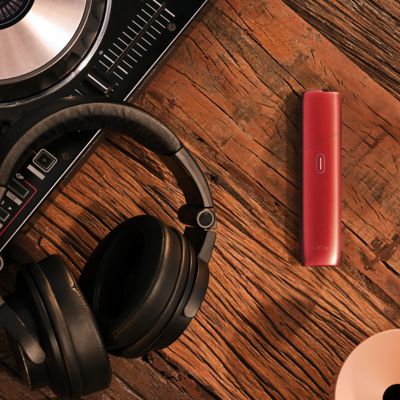 IQOS ORIGINALS ONE Scarlet neben DJ-Mischpult und Kopfhörern auf Holztisch.