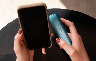 Hände halten IQOS ORIGINALS ONE Turquoise in der einen und Smartphone in der anderen Hand.