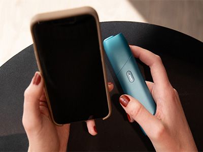 Person hält IQOS ORIGINALS ONE Turquoise in der rechten Hand und ein Smartphone in der linken.