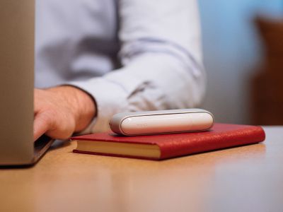 Ein weißes IQOS ORIGINALS DUO Gerät liegt neben einer Person, die an einem Laptop arbeitet.