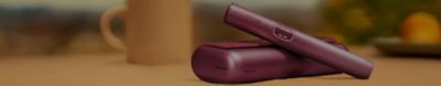 Stylish Garnet Red IQOS ILUMA i PRIME on a wooden table with a blurred cityscape background, next to a cozy armchair with an orange cushion.