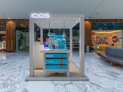 Vitrina frontal IQOS ILUMA con dispositivos exhibidos sobre base turquesa.