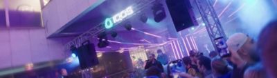 Dav ľudí pred pódiom s neonovým nápisom IQOS počas večerného eventu, pódiová konštrukcia so svetlami a reproduktormi, vystupujúca osoba s mikrofónom a návštevníci natáčajúci vystúpenie na mobilných telefónoch.