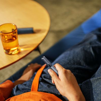 Zariadenie IQOS ILUMA v mužskej ruke, vedľa stolík s nabíjačkou a pohárom