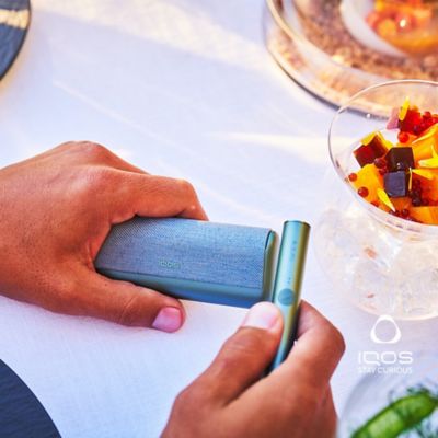 A person holding an IQOS ILUMA Prime holder & charger on the table