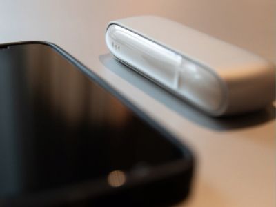 Ein weißes IQOS ORIGINALS DUO Gerät liegt neben einem Smartphone auf einer Oberfläche.