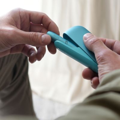 Zwei Hände bedienen das IQOS ORIGINALS DUO Turquoise vor hellem, unscharfem Hintergrund.