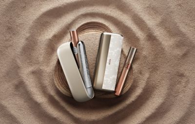 Dve napravi iz serije IQOS ILUMA i MINERA – ILUMA i ter ILUMA PRIME i – ter držala in žepna polnilnika na peščeni podlagi.