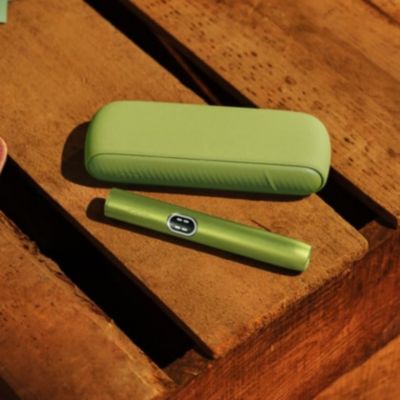 1 appareil IQOS ILUMA i mid couleurs Vert Anis posé sur un fond boisé.