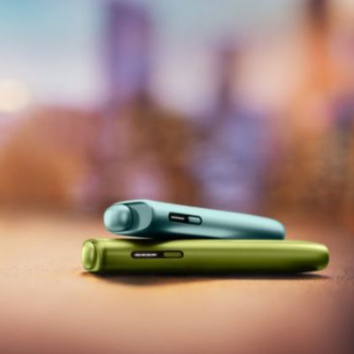 2 appareils IQOS ILUMA i ONE couleurs Vert Anis et Aigue-Marine posés l’un sur l’autre, sur un fond flouté. 