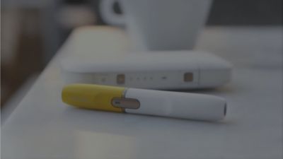 An IQOS 2. 4 plus lays on a white counter, next to it stands a coffee mug