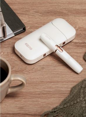 White IQOS 2.4 PLUS holder and charger on a wooden table next to a cup