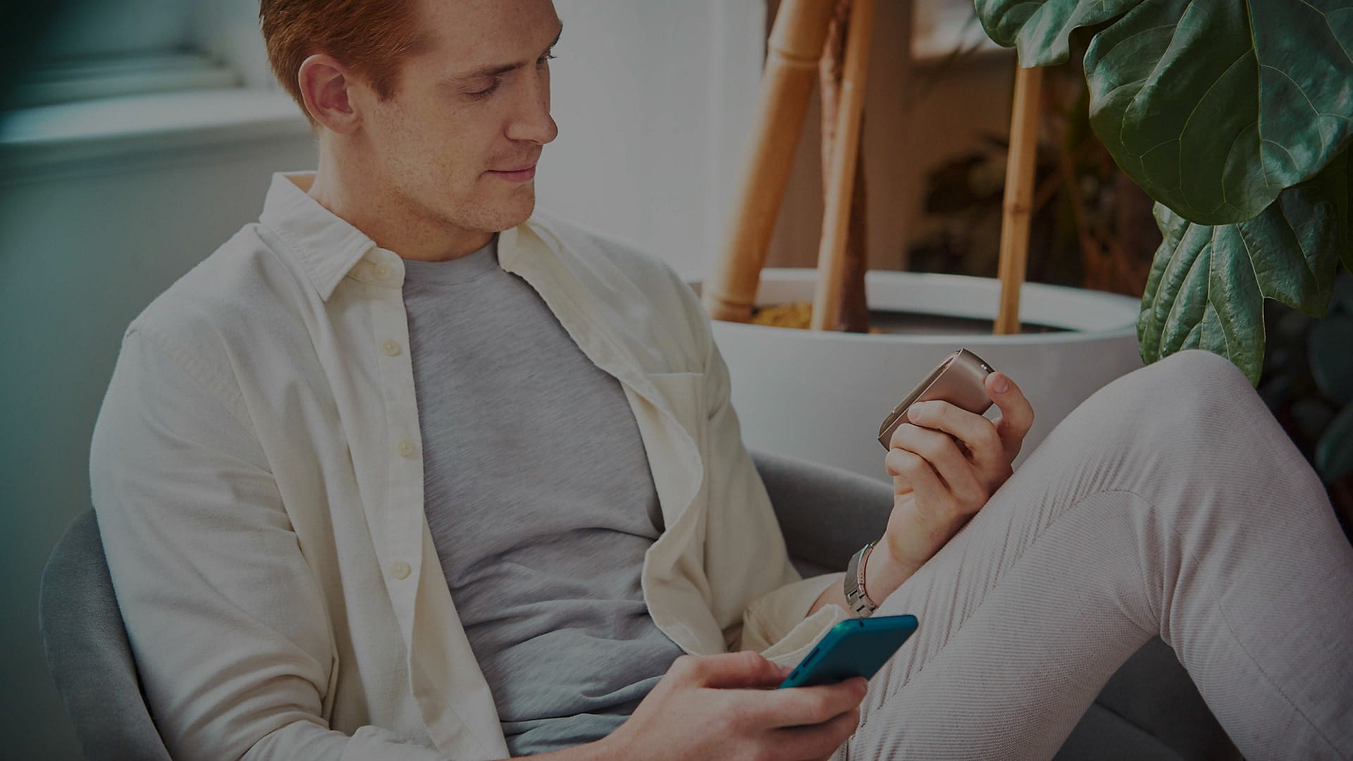 A man sitting in an armchair with his cellphone in his right hand looks to his left hand admiring his IQOS 3 DUO