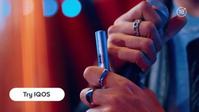 Close-up of hands holding an IQOS ILUMA i heated tobacco device with rings and blurred lights in the background.