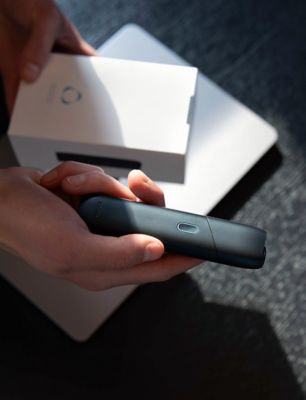Person hält IQOS ORIGINALS ONE Slate vor geöffneter weißer Geräte-Verpackung.