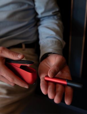 Person in hellblauem Hemd hält IQOS ORIGINALS DUO in Scarlet mit schwarzer Kappe in den Händen.