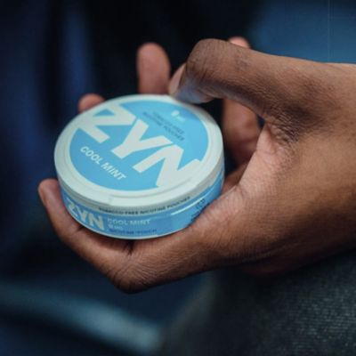 Hand holding a light blue ZYN Cool Mint can. Dark blue background.