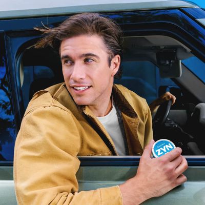 Muž v autě, dívá se z okna a drží modrou krabičku nikotinových sáčků ZYN Cool Mint