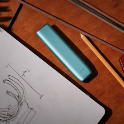 IQOS Iluma i turquesa colocado sobre boceto técnico en mesa de madera