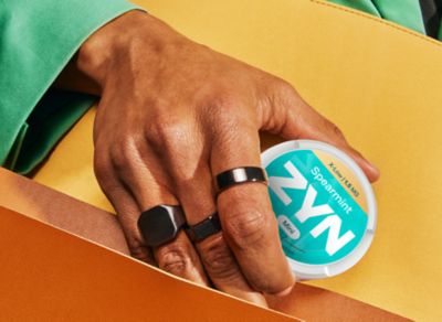 Ruka držící nikotinové sáčky ZYN na žlutém pozadí