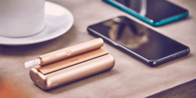 Ein IQOS ILUMA Prime in Gold neben Smartphones