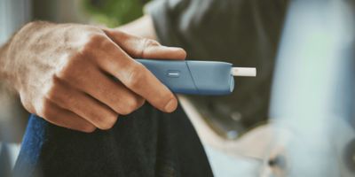 Ein Mann mit angewinkeltem Knie hält ein IQOS Originals One in der Hand.