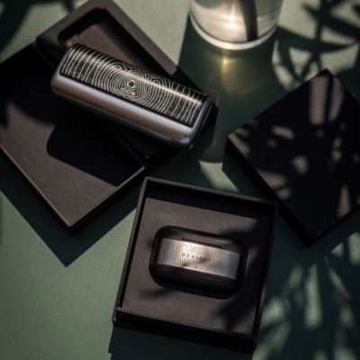 Set IQOS x DEVIALET zariadenia a slúchadiel v krabičke položený na stole v pozadí s pohárom vody.