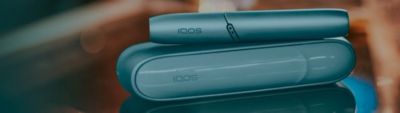 Zoom sur un charger IQOS avec un stylet IQOS posé dessus, eux-même posés sur une surement miroir avec en fond, un dégradé de couleurs orangées et bleues 