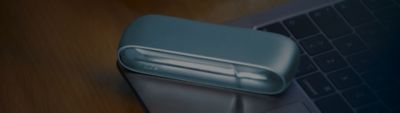 Image zoomée sur un IQOS 3 KIT posé sur le bord d’un clavier d’ordinateur portable, lui-même posé sur un bureau en bois 