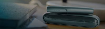 Zoom sur un charger IQOS avec un stylet IQOS posé dessus, eux même posés sur un bureau a coté de diverses fournitures de bureau. 
