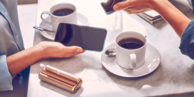 Händer von zwei Personen über einer Kaffeetiisch-Situation. Beide benutzen ein Smartphone und man sieht ein IQOS ILUMA Prime auf dem Tisch.