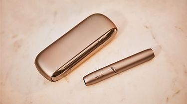 Gold IQOS DUO with a mobile, a glass of water and a yellow cup on a wooden table