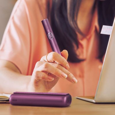 Frau hält IQOS ILUMA i Holder, ILUMA i Charger liegt auf Schreibtisch