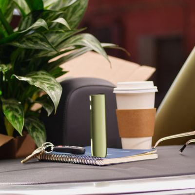 IQOS Iluma i ONE verde colocado sobre cuaderno junto a café y plantas en escritorio