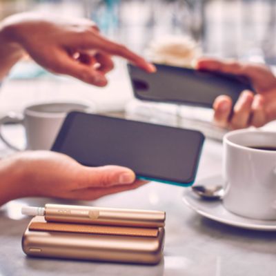 IQOS ILUMA Holder und Charger in Gold auf einem Tisch. Zwei Menschen tauschen sich zu ihren Smartphones aus.