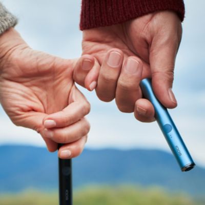 Zwei Personen haben sich am kleinen Finger untergehakt und halten jeweils ein IQOS ILUMA Device in der Hand