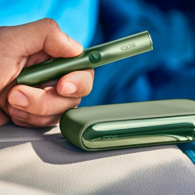 Jemand hält ein IQOS ILUMA PRIME Device in Moss Green in der Hand, darunter liegt ein Pocketcharger auf einer hellen Lederoberfläche