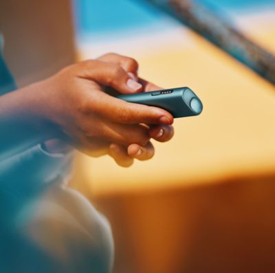 Mano sosteniendo un IQOS Iluma i ONE azul con fondo cálido desenfocado.