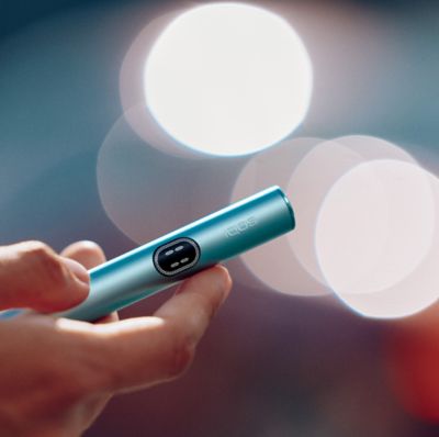 Mano usando un IQOS Iluma i ONE azul con luces difusas en el fondo.